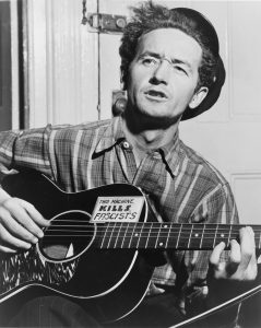 Woody Guthrie in 1943; the sticker on his guitar says “This Machine Kills Fascists”