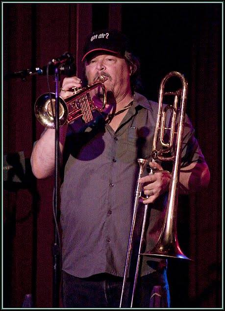 Mic Gillette (1951-2016) in a typical pose, holding his trumpet in his right hand and his trombone in his left